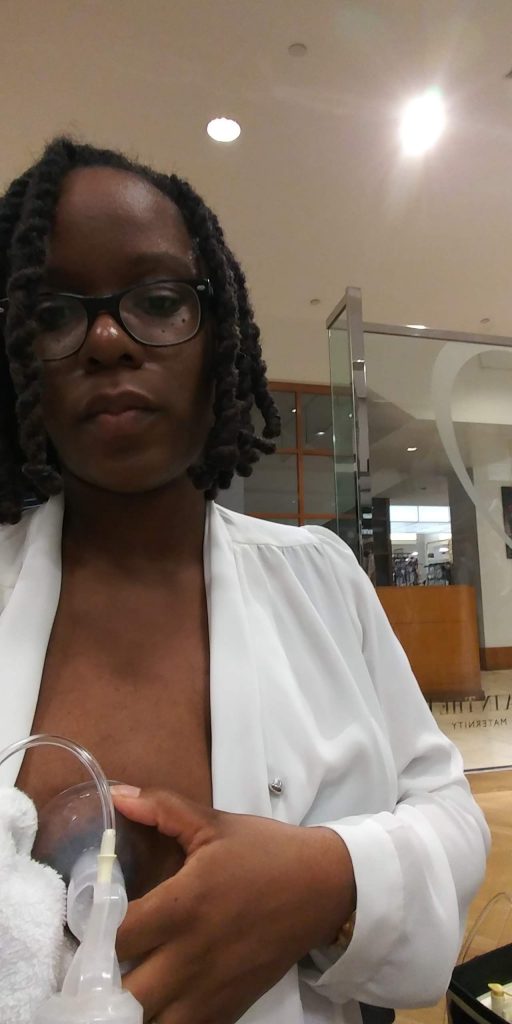 Black woman with dreadlocks wearing a long sleeve white blouse and glasses looks into the camera as she holds a flange to her breast.