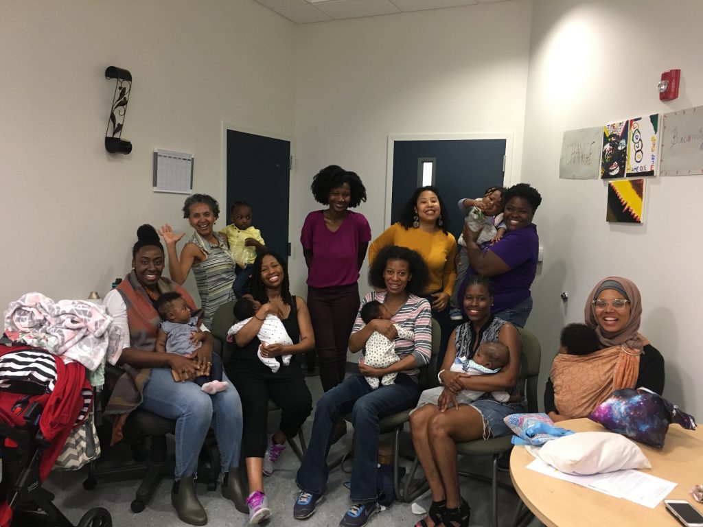 Group of Black women some sitting and some standing, most are holding babies and smiling.