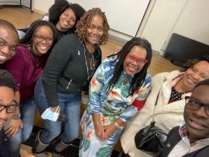 A selfie group picture of 8 people. The person to the far left is a Black man with a mustache, short black curly hair and is wearing black glasses. The person above him is a smiling Black man with black hair and a short buzzcut. The woman to the right is a smiling Black woman wearing a wine colored sweater, shoulder length black locs and burgundy glasses. The woman to the right and above the previous woman, is a smiling Black woman in a black t-shirt with thick black afro hair. The woman to the right of the previous woman is a smiling Black woman with light brown chin length locs, wearing a black long sleeved sweater, blue jeans and grey winter boots. The smiling Black woman to the right of the previous woman is slightly stooping down. She is wearing a floral dress, has armpit length thick black two-strand twists and red glasses. The woman to the right is a smiling Black woman with a cream colored winter coat and a black cross-body bag. The man on the far right is a smiling Black man with short black hair wearing a white collared shirt with blue and red color peaking out, a dark blue blazer and Black glasses
